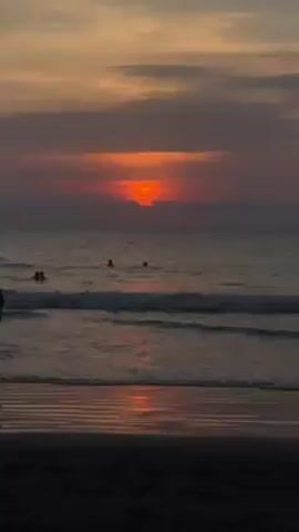 The video shows a beach at sunset. The sun is partially obscured by clouds, creating a reddish-orange glow. The ocean waves are small, and there are several people swimming in the water. The beach is dark, and the wet sand reflects the light from the sunset.