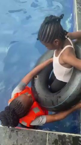 The video shows two young girls in a pool, one wearing a life vest and helping the other, who is in an inner tube, near the pool's edge. The camera pans up to reveal several people, mostly children, floating in inner tubes and life preservers in the pool. An adult male stands poolside, observing. The pool is surrounded by light-colored tiling and a building with a partially covered roof structure.  The sky is overcast.