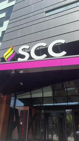 A short video shows the exterior of a building with the letters SCC prominently displayed above a bright pink band. A rainbow-colored flag is affixed to the left of the letters. Below the sign is a glass-covered entrance with a revolving door and several people, some carrying shopping bags. To the left of the entrance, a vertical sign reads "EXCHANGE" in red capital letters against a black background with orange geometric shapes. The building appears to be modern, with dark gray paneling and large windows reflecting the sky. The scene is outdoors on a sunny day, with a paved area in front of the building.