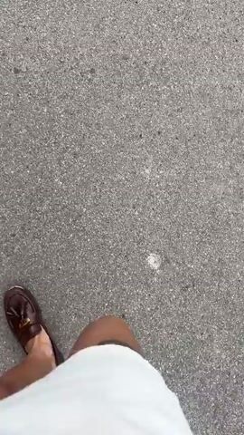 A person in a white garment and brown loafers walks on grey asphalt. A tattoo is visible above their knee. The camera is pointed down, providing a first-person view of the person's lower body and the ground. The person moves forward.