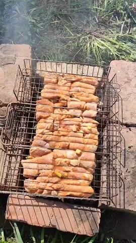 A close-up shot shows meat being grilled on a metal grate over a brick-built barbecue. The meat is arranged in rows and interspersed with pieces of onion or other vegetables. Smoke rises from the meat, indicating it is cooking. The barbecue is set on a ground covered with grass and some weeds. The camera slightly zooms in and out to showcase the meat and cooking process.