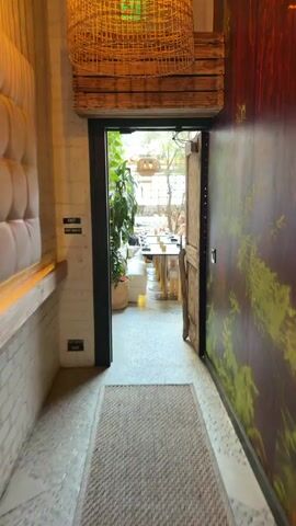 The video shows a point-of-view shot moving from a hallway into an outdoor dining area of a restaurant. The hallway is dimly lit with white brick walls and a wooden ceiling. There are exit signs on the left wall. The doorway leads to a bright, open-air space with several tables set for dining. Lush green plants hang from the ceiling and walls, and there are large wicker light fixtures. People are seated at some of the tables. The floor is tiled. The door on the right side of the frame is partially open and decorated with a green leafy pattern.