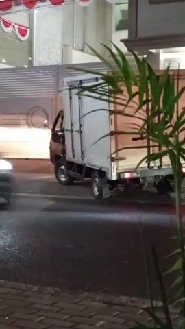 The video shows a black box truck parked on a street at night. There are red and white decorations hanging above a building in the background. A person is visible through the driver's side window of the truck. The scene is dimly lit, with lights from a nearby convenience store illuminating the area.