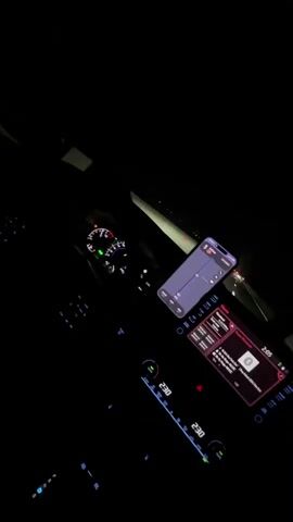 The video shows the interior of a car at night. The dashboard lights and screens are illuminated, providing a dim glow in the darkness. A smartphone is mounted on the dashboard, displaying what appears to be an audio equalizer interface. The car's central infotainment screen is also visible, showing music playback controls and song information. The speedometer and other gauges on the dashboard are lit up. The background outside the car is dark, with faint lights suggesting other vehicles or distant lights. The overall impression is a solitary drive at night with music playing.