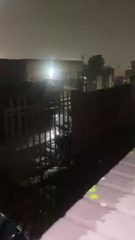 The video shows a nighttime scene, likely outdoors, with heavy rain. A metal gate is visible in the foreground, partially submerged in water that has flooded the area. Behind the gate, there are buildings and a streetlight that illuminates the wet surroundings. The video is shaky and seems to be taken from a slightly elevated position, possibly from inside a vehicle or under some form of cover. There are visible raindrops on the lens.