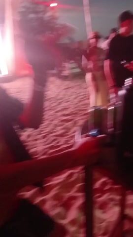 A person wearing a black and white striped shirt appears to be dancing on sand in an outdoor or partially covered event space. The lighting is primarily pink and the space seems to be set up with lounge furniture. The camera pans up to reveal a DJ booth with a screen displaying the text “Waikiki SEMANA SANTA 2023” along with palm tree graphics. Two DJs are visible in silhouette behind the equipment.