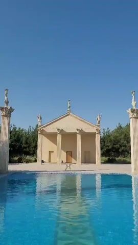 The video shows a view of a bright blue swimming pool with clear water. Behind the pool, there is a building with a classical architectural style, featuring columns and statues on its roof. The building has a triangular pediment and a set of double doors in the center. The entire scene is set against a clear blue sky and surrounded by green trees.