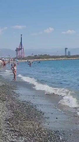 A person wearing a bikini stands on a pebble beach near the water's edge. In the background, several other people are visible in the water, some swimming and others on paddleboards. A large red and blue crane is prominent on the distant shore, along with buildings and mountains under a clear blue sky with scattered clouds. The waves gently lap at the shore.
