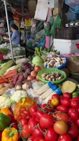 The video shows a stall at a market with a wide array of fresh vegetables and fruits. In the foreground there are several piles of tomatoes, and behind them there are pineapple halves, limes, mushrooms, cauliflower, sweet potatoes, carrots, bell peppers, cabbage, bananas, and other vegetables. A woman is seen behind the stall, weighing something on a scale. Plastic bags and other items hang from the top of the stall, and another woman is partially visible in the background. The scene is busy and colorful.