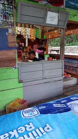 A young boy wearing a black t-shirt and shorts is at a small outdoor counter. He is handing a bag to a woman wearing a pink headscarf and a white and pink shirt. Behind them, shelves are stocked with various items. The counter is made of grey wood with drawers. To the right, there are rows of bottled water. The background shows trees and a stone wall. A blue tarp with text is in the foreground.