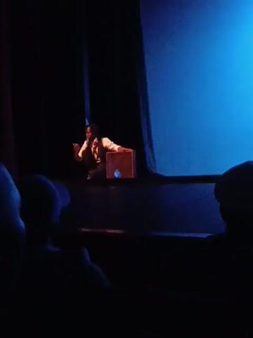 A person in formal attire, including a white shirt and dark vest, is on a dimly lit stage. They are crouched next to a rectangular wooden box with a blue emblem on its side. The person appears to be interacting with the box, possibly performing a trick or demonstration. A large, bright blue screen is visible to the right of the stage, creating a dramatic lighting effect. The audience is silhouetted in the foreground, their backs to the camera, indicating they are seated in a theater or performance space. The scene suggests an indoor event, likely a performance or show, taking place in Nairobi, Kenya. The mood is focused and perhaps a bit mysterious due to the lighting and the action of the performer.