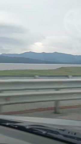 The video captures a view from inside a vehicle, likely a car, looking out at a landscape. The foreground shows a portion of the vehicle's interior, including the windshield and wipers. A silver guardrail runs horizontally across the middle of the frame, separating the road from the scenery beyond. In the background, a body of water, possibly a lake or river, stretches horizontally, bordered by a grassy field. Beyond the water, a range of blue-tinged mountains forms the horizon under a cloudy sky. A faint, circular logo, resembling a car manufacturer's emblem, is visible in the upper right corner of the sky. The overall impression is of a journey through a natural, scenic environment.