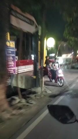 The video shows a view from a moving vehicle at night. A person on a red motorcycle is visible in the foreground, heading towards a large archway with illuminated signage. The archway has two large golden pillars and a white oval light fixture. Behind the archway, there are lights and buildings, including what appears to be a red and white striped barrier. The scene is blurry due to the motion of the vehicle and low light conditions.