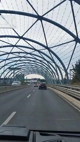 The video shows a first-person view from inside a vehicle driving on a highway under an overhead archway structure. The archway is made of metal beams and netting. Several cars are visible on the highway, driving in the same direction. On the sides of the highway, there are billboards and greenery. The sky is overcast.