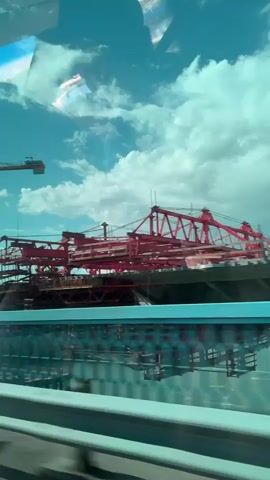 The video shows a view from a vehicle moving along a road, looking towards a large construction site. The primary focus is on a bridge under construction, characterized by its elevated structure and numerous cables. Towering cranes, one on the left and another red structure with multiple arms in the center, are visible, indicating ongoing construction work. The sky is bright blue with scattered white clouds. A blue barrier runs horizontally in the foreground, obscuring the lower part of the construction site. Reflections are visible on the window through which the video is recorded.