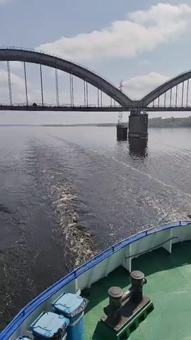 A high-angle shot shows a bridge in the distance from the stern of a boat. The bridge is a gray concrete structure, with tall curved arches supporting its roadway. The river water is a dark, calm surface, with visible ripples and foamy wake left by the boat. The boat’s edge is visible, painted white with blue trim, and is dotted with small, blue containers and black cleats. The sky is pale blue, sparsely covered with light clouds. The overall scene is one of steady movement and quiet water navigation under a clear sky.