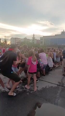 A group of people, mostly children, are playing tug-of-war with a thick rope. They are all on a paved surface, possibly a street or a courtyard. The sky is cloudy and the sun is setting, casting a warm glow. The people are dressed in casual summer clothing. The atmosphere appears to be festive and energetic.
