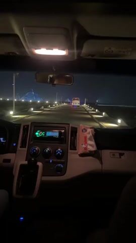 The video is taken from inside a vehicle, looking through the windshield. At night, the road ahead is lined with evenly spaced, square-shaped lights, leading towards the pyramids in the distance. Another vehicle, appearing to be a van or bus, is driving in the same direction, slightly ahead. Inside the car, the dashboard is visible, with a radio unit and various controls. A tissue pack is placed on the dashboard. Someone's hand can be seen briefly near the gear stick. The interior of the vehicle is relatively dark, with the overhead light on.