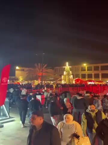The video captures a nighttime scene of a public gathering, possibly a Christmas market, illuminated with colorful lights and laser beams. A large, cone-shaped Christmas tree stands prominently in the background, adorned with bright lights. People of various ages and ethnicities are seen strolling around, some pausing to chat, while others appear to be filming or taking pictures with their phones. There are stalls or vendor booths under red umbrellas, suggesting a market atmosphere. A red barrier surrounds a particular area, possibly delineating a specific activity or event space. The general mood appears festive and lively.