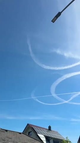 A vertical video shows a clear blue sky with several circular contrails left by aircraft. The sun is visible and shining brightly. In the foreground, the tops of houses with solar panels and trees are visible. A street lamp extends into the frame from the top right corner. The contrails form overlapping circles and lines, creating a dynamic pattern against the sky. The overall impression is of a sunny day with active air traffic.