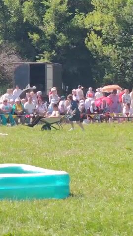 A group of spectators sits on a grassy hill watching a competition. Two young people are participating in an activity with a wheelbarrow. One person is pushing the wheelbarrow, which has another person sitting in it. The person in the wheelbarrow appears to be holding a long stick. They are in a field with trees in the background and a small blue inflatable pool in the foreground. The weather appears to be sunny and warm.