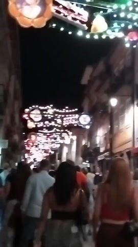 The video shows a street at night decorated with numerous lights strung across it, likely for a festival or celebration. A crowd of people are walking down the street, viewed from behind. The people are dressed in casual clothing. The street appears to be lined with buildings. The lighting is dim, with the primary illumination coming from the decorative lights and some streetlights. The scene suggests a lively, social atmosphere.