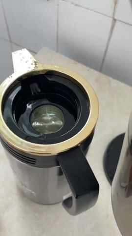 The video shows a close-up of someone pouring clear, steaming water from a black kettle into a silver and gold insulated carafe with a black handle. The carafe sits on a white countertop, and white wall tiles are visible in the background. The kettle is held out of frame. The focus is on the act of pouring the water.
