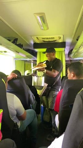 The video shows a man selling food on a bus. The man, wearing a black t-shirt, ripped jeans, and a black baseball cap, is smiling and holding a stack of what appears to be fried plantain sandwiches and a metal pot. He is moving through the aisle of a bus, which has multiple passengers seated. The interior of the bus is green, with overhead storage compartments. The man seems to be interacting with the passengers, offering the food for sale.