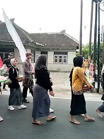 A group of people are walking in a parade. Most of them are carrying flags of various colors, including red, white, orange, yellow, and blue. Some of the people are wearing traditional clothing, including headscarves and patterned sarongs. One person in the front is carrying a basket of what appears to be food. The parade is taking place on a paved street, with houses visible in the background. The sky is overcast.