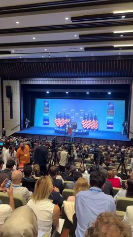 The video shows a stage with a large screen displaying the NATO logo and text that appears to read "NATO OTAN ANKARA". In front of the screen, there are several rows of American flags. On a raised platform in the center of the stage, three men are gathered around a podium. One man, wearing a dark suit, is speaking or gesturing at the podium. Two other men in suits are standing near him. One of these men appears to be adjusting something on the podium. Another man in a blue suit is walking away from the podium towards the right side of the stage. The stage is set up in a large auditorium or conference hall, filled with an audience seated in rows of chairs. Many individuals in the audience are holding up mobile phones, possibly to record or take pictures. Numerous tripods equipped with cameras are positioned in front of the audience, indicating a press event. The overall atmosphere suggests a formal gathering, likely a press conference or a formal ceremony.