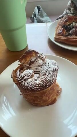 A close-up, top-down shot shows a white plate with a golden-brown pastry dusted with powdered sugar. To the left of the plate is a portion of a light green cup. In the background, another white plate holds a rectangular pastry with a dark filling. A wooden surface is visible beneath the plates and cup. The scene appears to be indoors, possibly a cafe, with natural light filtering in from a window behind the cup.