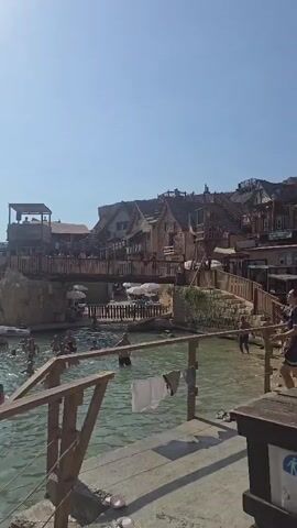 The video shows a group of people swimming and playing in the ocean on a sunny day. There are several boats in the water, and a building perched on a cliff overlooking the scene. The water is clear and blue, and the sky is clear and bright. Some people are splashing and jumping in the water, while others are swimming or relaxing on the boats. The overall atmosphere is one of fun and enjoyment.