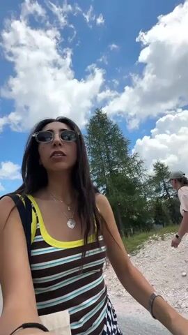 A woman wearing sunglasses and a striped tank top walks down a paved path next to a grassy hill. She is looking up at the sky, which is blue with white clouds. In the background, a large rock formation towers over a treed landscape. Another woman with a backpack walks away from the camera down the same path. Further in the distance, mountains are visible under the cloudy sky. A white van and another vehicle are parked on the side of the road.