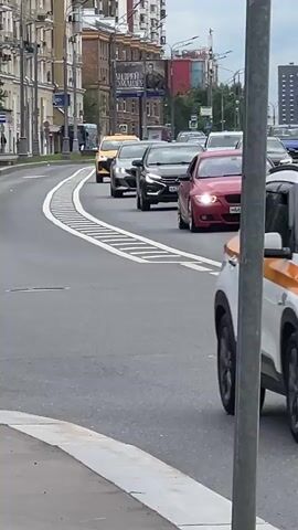 A first-person perspective video captures a street scene with several cars driving on a multi-lane road. The road has white markings, including a dashed line and a solid white line, indicating lanes. On the left side of the road, there are tall buildings with windows and balconies, as well as streetlights and trees. On the right side, there's a gray pole in the foreground. The cars visible are a yellow taxi in the foreground on the left, a black car and a silver car in the middle lanes, and another yellow taxi further ahead on the right. The traffic appears to be moving. The sky is overcast.