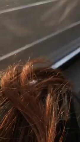 The video shows a close-up view of a person with reddish-brown hair. The camera angle is low, looking up at the person. The background is blurry and appears to be the interior of a car, with a roof and part of a window visible.