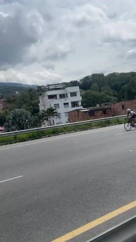The video shows a cyclist riding along a road. The cyclist is wearing a helmet, a white shirt, and dark shorts, and is riding a black road bike. The road is asphalt with a yellow line marking the edge. There is a metal guardrail on the side of the road. In the background, there is a town with houses and trees, and mountains in the distance. The sky is cloudy. As the cyclist approaches the end of the road, there is a group of people and some uniformed individuals standing on the side. There is also a pedestrian overpass in the background.