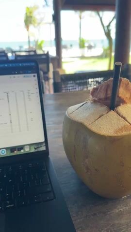 A laptop sits open on a weathered wooden table, displaying a spreadsheet with columns labeled "Google Ads," "Profile Data," and "Training." Next to the laptop is an opened green coconut, its top cut to reveal the white flesh inside. The setting is a covered patio or veranda, offering a clear view of a sunny beach with the ocean in the background. There are empty wooden tables with chairs arranged outside, giving the impression of a relaxed, possibly beachside cafe or restaurant. The angle of the camera is low, looking up and out towards the beach view. Above the laptop and table, the roof is visible, constructed with rustic materials. There is no indication of any people present in the immediate frame.