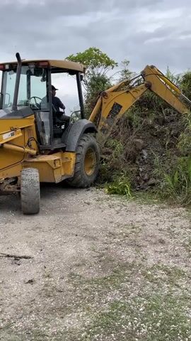 A yellow backhoe is operated by a person sitting in the driver's seat. The backhoe's arm is extended and digging into a pile of dirt, rocks, and vegetation. The scene appears to be outdoors, possibly in a rural or undeveloped area, with a cloudy sky and trees in the background. The ground is covered in gravel.
