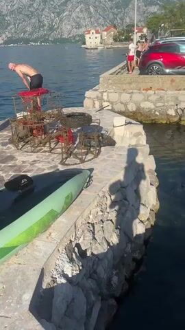A bald man with a beard and tattoos, wearing black shorts, stands on a stone pier next to a body of water, looking down at several rusty metal cages and a discarded tire, all covered in marine growth, which are placed on the pier. A green and black paddleboard lies on the pier nearby. Further along the pier, a red SUV is parked, and several other people are visible near the water. In the background, mountains rise across the water, and a white building with a red roof is visible on the distant shore. The scene is illuminated by bright sunlight, casting long shadows on the pier.