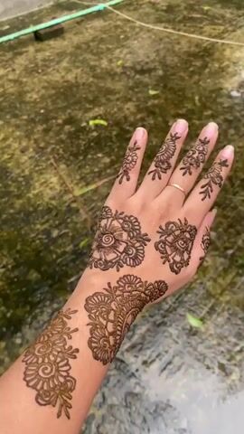 The video shows a woman's hand adorned with henna tattoos. The tattoos feature intricate floral designs on her fingers, the back of her hand, and extending onto her wrist. She wears a gold ring on her ring finger. The background varies, showing a wet, patterned surface, then shifting to a green, tree-filled area with a concrete wall. The hand is held up and rotated to showcase the henna designs from different angles.