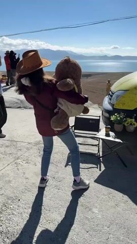 The video captures an outdoor scene featuring a person holding a large teddy bear against a stunning natural backdrop. The person, seen from behind, wears a brown cowboy hat, a maroon long-sleeved top, blue jeans, and athletic shoes. They are holding a substantial brown teddy bear, which appears to be wearing a small jacket, and are positioned facing a vast body of water, likely a lake, with distant mountains under a clear blue sky.

To the right of the frame, a yellow van is parked, possibly a camper van or mobile vendor. The van's side displays Chinese characters along with some partially visible English text that seems to read "I Live Lo..." or "I Love Lo...". In front of the van, a small black folding table is set up, adorned with several small potted plants, including sunflowers. Two small chairs, one black and one pink, are placed near the table.

On the far left, another individual is partially visible, holding a smartphone up, suggesting they are taking a photograph of the person with the teddy bear. The ground is a paved surface. The strong shadows cast by the subjects indicate bright sunlight, suggesting it's either mid-morning or afternoon. The setting appears to be a scenic overlook in Gyirong County, China.