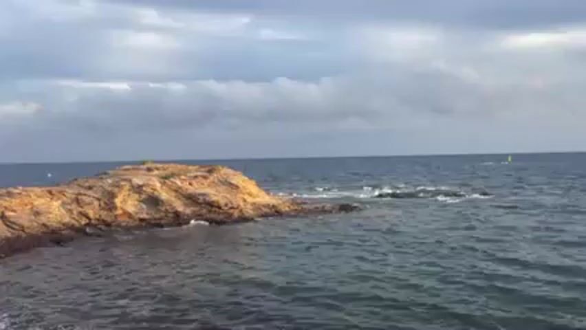 The video shows a rocky outcropping jutting into a body of water. The rocks are predominantly brown and yellow. The water is choppy and dark blue. The sky is overcast with gray and white clouds. A small yellow buoy bobs in the distance. There are no visible people or specific actions occurring.