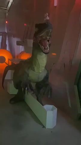 This is a close-up video of a dinosaur animatronic, likely a velociraptor, in a dimly lit indoor setting. The animatronic is green and brown with sharp claws and teeth, its mouth open in a roar. It appears to be standing on some sort of platform or prop, possibly part of an exhibit or attraction. The background shows metallic walls and structures, suggesting a science fiction or theme park environment. Red and green lights are visible in the background, adding to the atmospheric lighting. The camera is shaky at times, and the focus shifts slightly, but the dinosaur remains the central subject. There are no people visible in the video, and the focus is solely on the animatronic dinosaur.