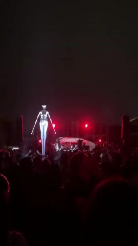 A drone shaped like a humanoid figure with a reflective surface is illuminated by red and white lights. It hovers above a crowd of people who are looking up at it. The crowd is gathered in an outdoor space at night. There are structures in the background that appear to be part of a stage or lighting rig. The drone moves slightly, and the crowd reacts with excitement.