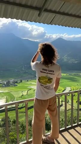 A man with curly brown hair stands on a balcony, looking out at a valley of rice paddies. He wears a white t-shirt and light brown pants. The shirt has a design on the back that says "Mao Sapa Trekking and Homestay." The valley is filled with green rice paddies, some of which are flooded with water, reflecting the sunlight. Mountains rise in the distance.