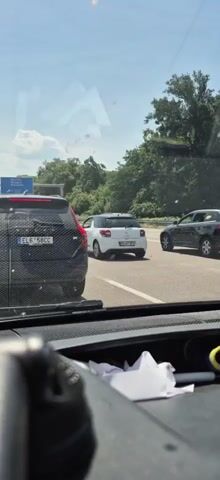 The video shows a view from inside a car looking out at a traffic jam on a highway. Several cars are visible, including a black SUV in the foreground on the left, a white compact car in the middle, and a black SUV on the right. Another dark-colored SUV is visible behind the white car. The sky is bright and cloudy. Trees and vegetation line the side of the highway. The dashboard of the car from which the video is being filmed is visible in the foreground.