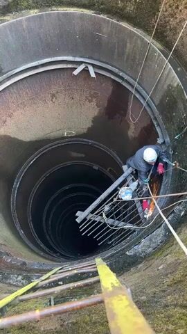 A person wearing a white hard hat and a grey jacket is working inside a large, cylindrical well. They are attached to ropes for safety and are using a grinder to cut metal. Sparks are flying from the grinder. A metal ladder is visible at the bottom of the well, and another ladder or scaffolding is positioned on the side. The well walls appear to be made of concrete and metal, with signs of rust. The overall environment is industrial and suggests maintenance or repair work is underway.