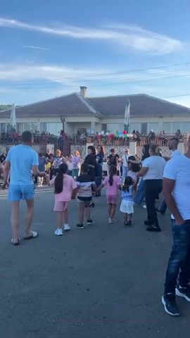 The video shows a group of people dancing in a street in front of a building. The building appears to be a restaurant or a similar establishment, with outdoor seating and a patio. The sky is blue with some clouds, and the sun is shining, suggesting it is daytime. The people are of various ages, and some are children. They are holding hands and dancing in a circle or in groups. A woman in a black dress appears to be singing or performing. There are some decorations, including balloons, visible on the patio of the building. The overall atmosphere seems festive and celebratory.