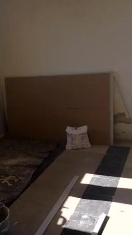 A person stands in a room with a window. The room is in various stages of renovation, with materials on the floor, including a bucket, a ladder, and a bag. The walls are partially finished, and the floor is covered in a protective sheet. A door with a decorative pattern is visible in the background. A large wooden cross hangs on the wall, and a television is mounted above it. There are chairs in the room, and a speaker is placed on a raised platform.