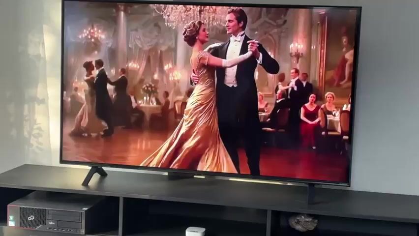 The video shows a large television screen displaying a scene from a ballroom dance. Two people, a man and a woman, are dancing in the foreground. The woman is wearing a long, flowing gold dress, and the man is wearing a tuxedo. In the background, other couples are dancing, and there are tables set with food and drinks. The overall scene suggests a formal event or party. The television is placed on a black stand in front of a grey wall. To the left of the television, there is a black computer tower on a shelf.